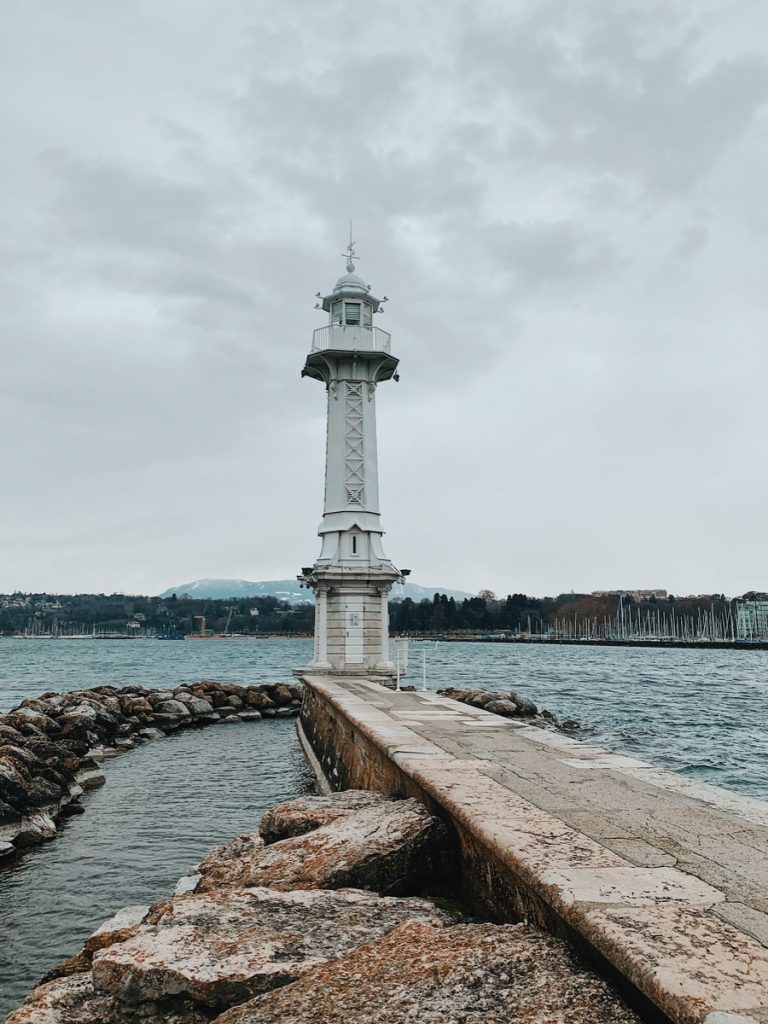 white lighthouse near body of water viewing buildings under white sky